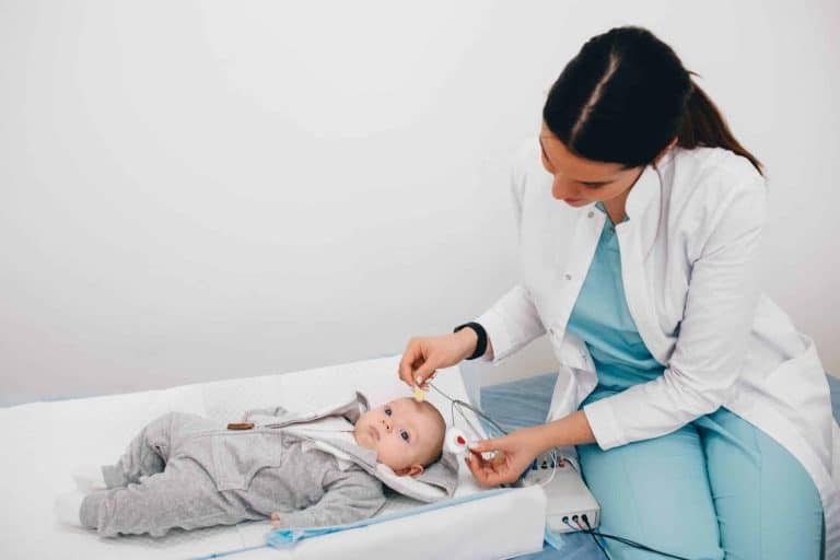 Pediatric Hearing Screening A baby in a onesie lying on a bed while a doctor places electrodes on it's head.