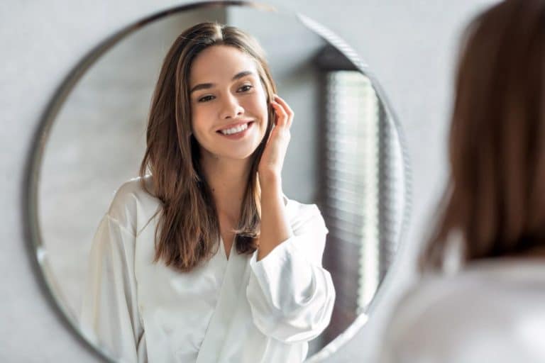 After a Chemical Peel Woman with brunette hair and eyes looking into a bathroom mirror and smiling at herself. One hand is up against her cheek, lightly touching her long hair. The other hand is at her side and out of frame.