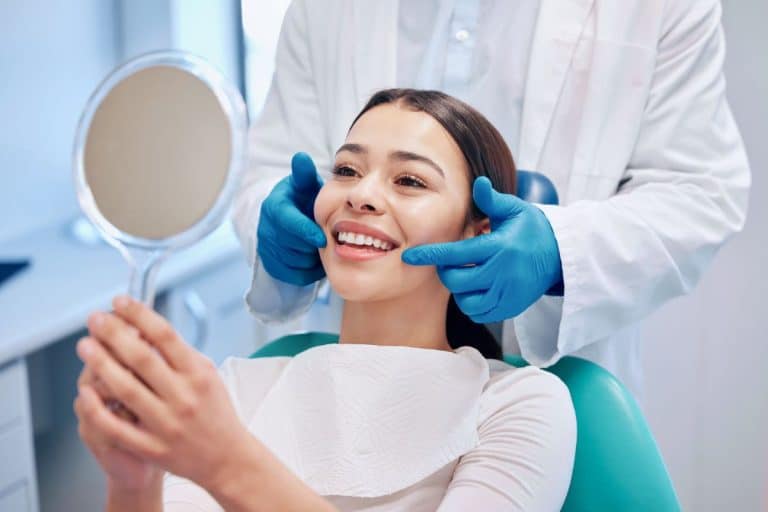 Before Face Lift Consultation A woman in a dental style chair looks in a mirror and smiles. A doctor is behind her using gloved hands to point at her checks with gloved hands.