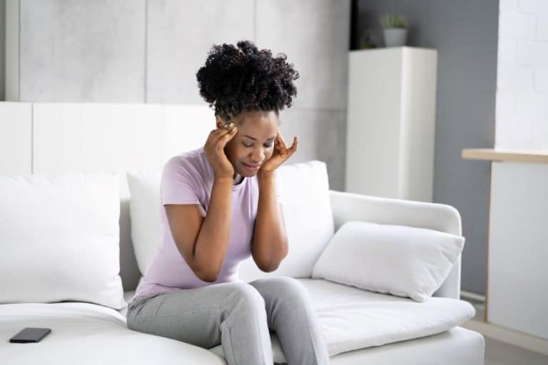 suffering from Benign paroxysmal positional vertigo Woman on a couch in a modern living room with her hands against her temples and her head turned downwards. Her facial expression is neutral, but her eyes are closed.